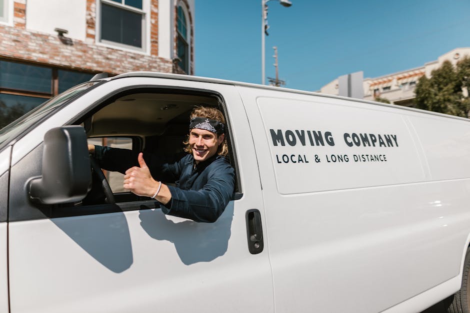A person wearing a beige jacket and black cap is seen loading a cardboard box into the rear of a white van parked on a residential street, part of a house removal process. The van, positioned beside a