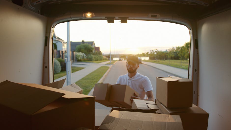 A smiling man with long hair, wearing a bandana and a dark jacket, is seated in the driver's seat of a white moving van with the words 'Moving Company Local & Long Distance' printed on the side. He is giving a thumbs-up gesture through the open window. The van is parked outside a building on a street, with a clear blue sky overhead. Visible objects inside the van include cardboard boxes, wrapped furniture, and packing materials, indicating preparations for a home relocation. The loading process involves the vehicle being ready for furniture transport and packing and moving activities, with the environment featuring typical urban surroundings and the van positioned on the pavement.